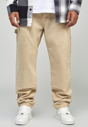 Beige cargo pants with curved side pockets, a relaxed fit, and ribbed cuffs, paired with white sneakers and a black and white flannel shirt.