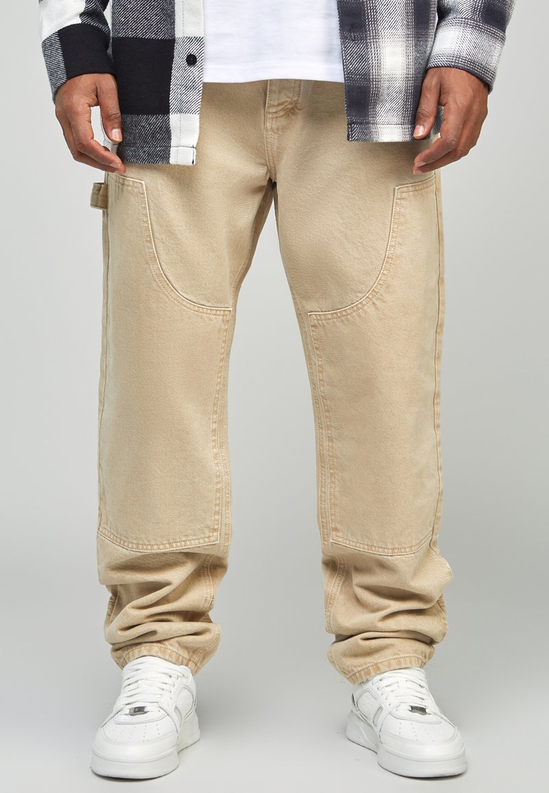 Pantaloni cargo beige con tasche laterali curve, una vestibilità rilassata e polsini a coste, abbinati a sneaker bianche e una camicia a quadri bianca e nera.