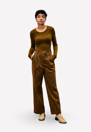 Femme debout avec les mains dans les larges poches de son pantalon marron, portant un haut ajusté à manches longues olive et des mocassins à talons beiges sur fond blanc.