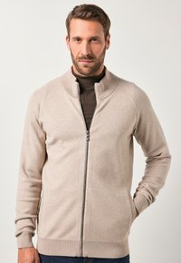 Cardigan zip a leggera beige con colletto e polsini a coste, caratterizzato da una texture liscia e tasche laterali, indossato sopra una maglia scura.