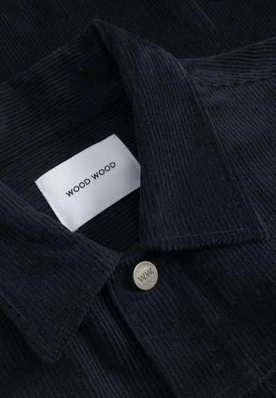 Černý kordový límec košile s etiketou s nápisem "WOOD WOOD" v černém písmu a stříbrným knoflíkem s vygravírovaným "W.W.".
