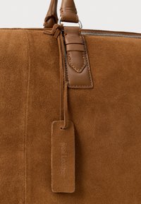 Bruin suède leren tas met een gestikte leren handgreep en een suède leren label dat bevestigd is nabij de metalen ritssluiting.