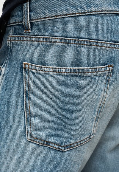 Gros plan sur la poche arrière d’un jean en denim bleu avec des coutures orange visibles et une boucle de ceinture au-dessus de la poche.