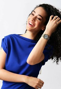 Orologio da polso argento con quadrante rotondo e dettagli grigi. Il modello indossa una maglietta blu vivace con una texture liscia, con drappeggio sulle spalle e maniche corte.