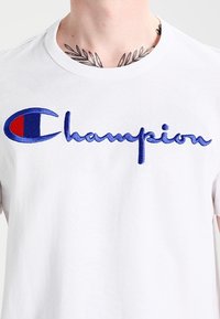 Camiseta de algodón blanca con gran logo bordado en azul de "Champion" con acento rojo en la manga izquierda. Cuello redondo y mangas cortas.