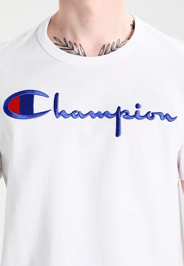 Camiseta de algodón blanca con gran logo bordado en azul de "Champion" con acento rojo en la manga izquierda. Cuello redondo y mangas cortas.