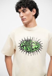 Tricou supradimensionat din bumbac crem, cu un design grafic central în verde și negru, având cuvântul "ARIES" și accente de raze solare.