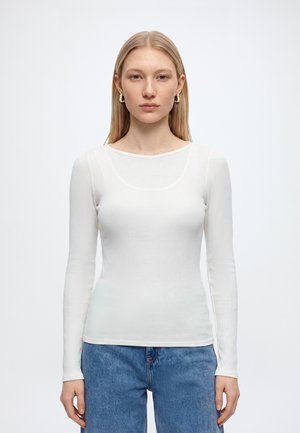 Jeune femme aux cheveux blonds et raides porte un haut blanc à manches longues côtelé, un jean bleu et des boucles d’oreilles créoles dorées, face à nous avec une expression neutre.