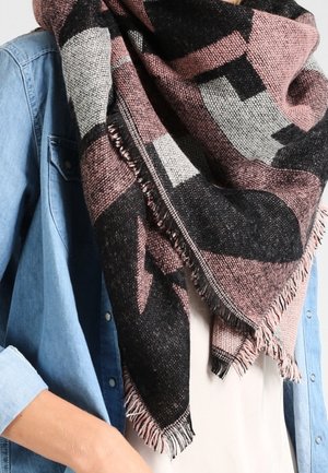 Personne portant une grande écharpe triangulaire tissée aux motifs géométriques noirs, roses et gris, sur une chemise en denim bleu clair et un haut blanc.