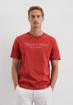 Crvena pamučna majica s kratkim rukavima, s natpisom "Marc O’Polo EST. U STOCKHOLMU" otisnutim svijetlim slovima preko prsa. Glatka tekstura.