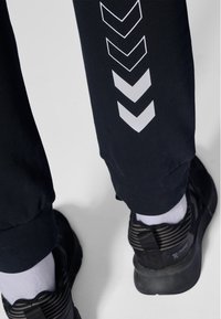 Czarne spodnie dresowe z białymi wzorami w kształcie szewronu na nogawce; połączone z czarnymi butami sportowymi i białymi skarpetkami widocznymi przy kostce.
