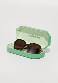 Occhiali da sole con lenti marroni scure e montatura nera spessa, esposti all'interno di un astuccio ottagonale verde con la scritta "SWAROVSKI" stampata all'interno.
