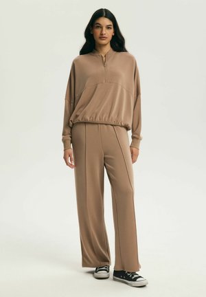 Žena oblečená v voľnom béžovom topu na zips a zodpovedajúcich širokých nohaviciach, v čierno-bielych teniskách, stojaca pred jednoduchým pozadím.