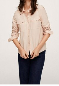 Chemise bouton-up rose clair avec des manches retroussées, deux poches poitrine et une texture douce, associée à un jean en denim bleu foncé.
