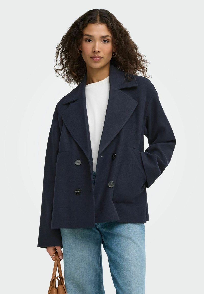 Femme aux cheveux bouclés portant un manteau bleu marine à double boutonnage, un haut blanc, un jean bleu clair et tenant un petit sac à main marron.
