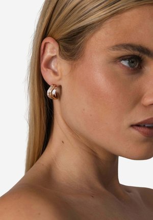 Gold- und weiße Ohrringe mit einem verdrehten Design und kleinen kreisförmigen Akzenten. Die Ohrringe werden auf einem hellen Hautton getragen.