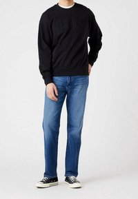 Felpa nera con la scritta "WRANGLER" in rilievo sul petto, abbinata a jeans blu e sneakers nere, in piedi davanti a uno sfondo neutro.