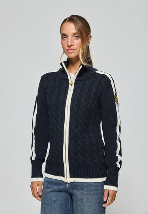 Navy knitted cardigan with cable pattern, white accent stripes, high collar, and gold zipper. Features an embroidered badge on the sleeve.