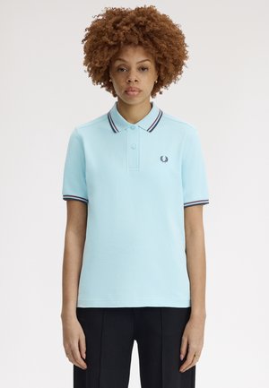 Mujer con cabello rizado, vestida con una camisa tipo polo azul claro con cuello a rayas y pantalones negros, de pie y recta contra un fondo liso.