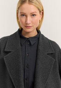 Blonde Frau mit blauen Augen, die goldene Creolen trägt, ein dunkles, geknöpftes Hemd und einen schiefergrauen Mantel mit breiten Revers.