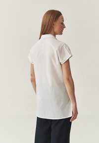Bílá košile s krátkým rukávem, volným střihem, límcem a rovnými lemy. Vyrobena z lehké tkaniny, hladké struktury a s minimálními designovými detaily.