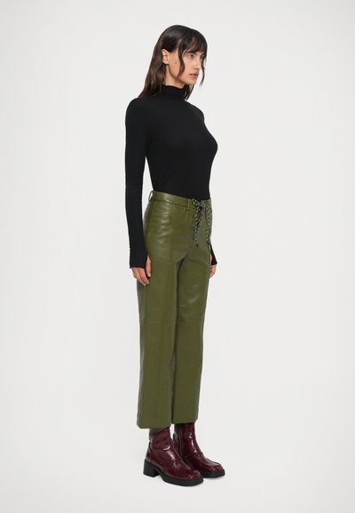 Blusa preta de mangas compridas combinada com calças de imitação em pele verde-oliva de cintura alta, com frente amarrada e botas de tornozelo burgundy brilhantes.