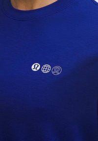 Kék ing, három kis fehér logóval a mellkason: két Lululemon szimbólum és egy földgömb ikon közöttük.