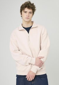 Sweat-shirt rose clair avec un col montant et une fermeture éclair quart. Tissu doux, coupe décontractée et poignets côtelés. Porté par-dessus un t-shirt bleu marine et un jean.