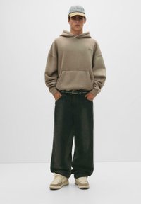 Beige oversized hoodie med en frontlomme, sorte vidde jeans og cremefarvede sneakers. Lys blå kasket med logo accent, der har en mørk skygge.
