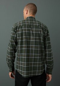 Chemise en flanelle à carreaux verts avec des accents gris et jaunes. Manches longues, poignets boutonnés et ourlet arrondi. Le matériau semble doux et texturé.