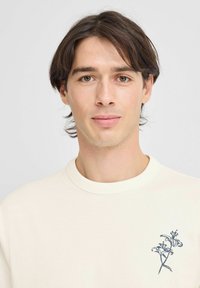 Cremefarvet t-shirt med rund hals, lavet af bomuld, med et blåt blomsterdesign på venstre brystområde, glat tekstur og afslappet pasform.