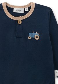 Marineblaues Langarmhemd mit beigem Kragen, das auf der Brust einen gestickten blauen Traktor und einen Holzknopf am Hals aufweist.