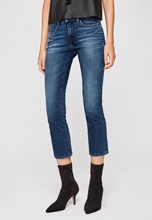 Nainen pukeutuneena lyhytlahkeisiin sinisiin farkkuihin ja mustiin kärjellisiin nilkkabootseihin, joissa on keskipitkät korot, valkoista taustaa vasten.