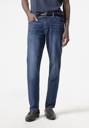 Hombre vestido con vaqueros azules ajustados, un cinturón negro, una camisa oscura metida por dentro y zapatos negros de cuero sin cordones, de pie con una mano en el bolsillo.