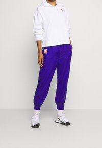 Bílá mikina s kapucí a přední kapsou, a fialové tepláky s elastickými manžetami. Pozoruhodné detaily zahrnují značkové nášivky na kalhotách a botách.