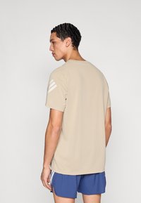 Camiseta beige de algodón con mangas cortas, cuello redondo y tres rayas blancas en el hombro izquierdo, combinada con pantalones cortos azules.