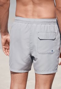 Grijze zwemshorts met een elastische tailleband, voorzien van een achterzak met klep en een decoratief blauw oogje. Gemaakt van lichtgewicht materiaal.
