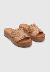 Sandalias de plataforma color beige con una tira ancha que presenta tachuelas doradas. La suela es más gruesa y está contorneada para brindar soporte, ofreciendo un diseño casual.