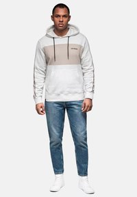 Sudadera gris claro con franja horizontal beige y cordones negros, combinada con jeans azules y zapatillas blancas. Presenta un bolsillo frontal y un logo.