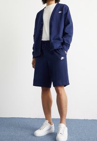 Námořnický modrý bavlněný kardigan s knoflíkovým uzávěrem a logem Nike, doplněný o odpovídající šortky. Bílé tenisky dotvářejí outfit.