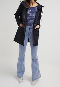 Marinblå huva jacka med dragsko, blå blommig grafisk t-shirt, ljusblå utsvängda jeans och grå sneakers med en texturerad yta.