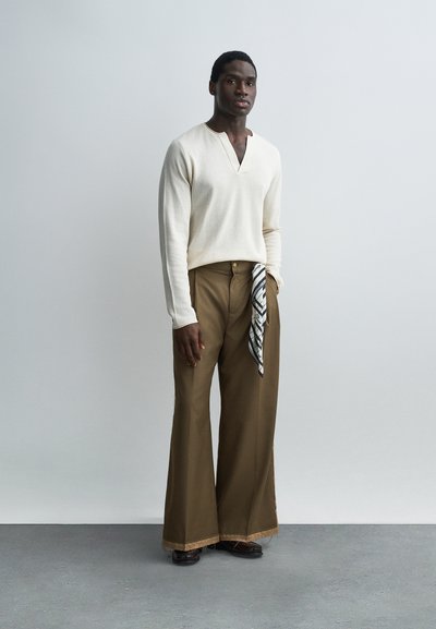 Uomo in piedi contro un muro semplice che indossa un maglione crema con scollo a V, pantaloni marroni a gamba larga, scarpe scure e un panno decorato infilato nella tasca.