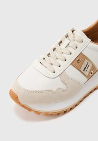 Baskets blanches et beiges avec semelle en caoutchouc, détails en daim beige, lacets beiges, et étiquette latérale indiquant "Blauer U.S.A."