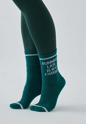 Nogi w ciemnozielonych legginsach, noszące turkusowe prążkowane skarpetki z białymi paskami i napisem „RUNNING LATE IS MY CARDIO” na jasnym tle.