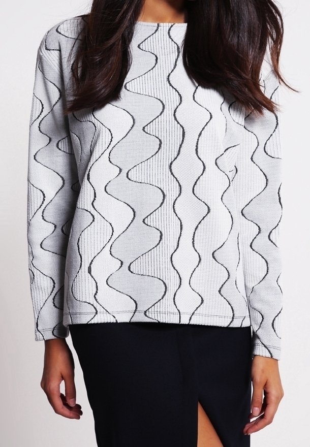 Femme portant un pull blanc à manches longues avec des motifs de lignes ondulées noires et une jupe noire avec une fente à l'avant.