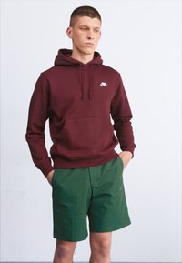 Sweater com capuz marrom escuro com bolso frontal e logotipo branco, combinado com calções verdes. Ambas as peças têm uma textura suave e um corte descontraído.