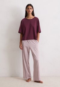 Vrouw die blootsvoets staat in een losse bordeauxrode top met korte mouwen en lichtgekleurde, gestreepte wijde broek tegen een effen muur.