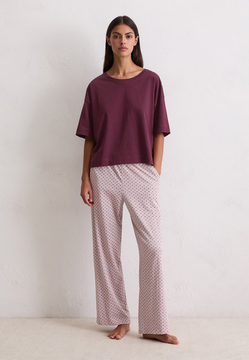 Vrouw die blootsvoets staat in een losse bordeauxrode top met korte mouwen en lichtgekleurde, gestreepte wijde broek tegen een effen muur.