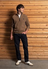 Sudadera marrón con cremallera, jeans oscuros y zapatillas blancas con detalles en negro. De pie contra una pared de madera con tablones horizontales.