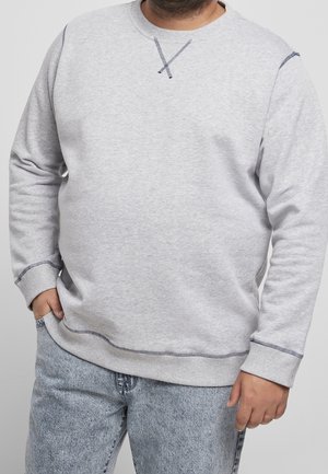 Homme portant un sweat-shirt gris clair avec des surpiqûres foncées et un jean bleu clair, une main dans la poche, debout devant un fond uni.